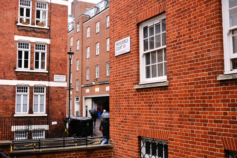 The image shows a narrow alleyway between two red brick residential buildings in an urban setting. On the right side, the building features a large, white-framed sash window with multiple panes and a stone sill. Below, there is a smaller window with a decorative black iron grille. The building on the left also has multiple sash windows with white frames and small flower boxes on some window sills, adding a touch of greenery. In the background, a black street lamp is visible mounted on the side of a building. At ground level, two black waste bins with yellow and green markings are placed near a metal railing and pavement, partially obscured by a person dressed in casual clothing, including a jacket and jeans, who appears to be walking away. The alleyway is intended for private waste collection, and Waste Disposal Marylebone could provide such rubbish removal services in similar settings, supporting alternative waste handling outside of municipal collection systems.