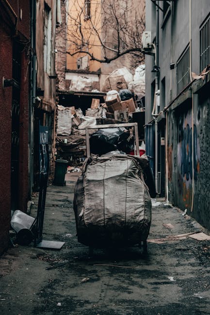 A narrow urban alleyway filled with miscellaneous household rubbish and debris, including large black garbage bags and cardboard boxes, stacked against a wall at the far end. The foreground features a metal shopping trolley covered with a worn, dirty fabric bag, positioned centrally on the asphalt ground which appears stained and littered with small scraps. On the left side, a small black waste bin is tipped over, with some loose rubbish nearby. The alley is flanked by tall buildings with graffiti and peeling paint, and a leafless tree is visible behind the mound of waste, indicating an outdoor environment. Fluorescent lighting casts a muted, dull atmosphere over the scene. The cluttered scene exemplifies a typical site for private rubbish collection or on-site clearance, hinting at a service such as Waste Disposal Marylebone’s rubbish removal operations that manage urban waste outside municipal collection areas.