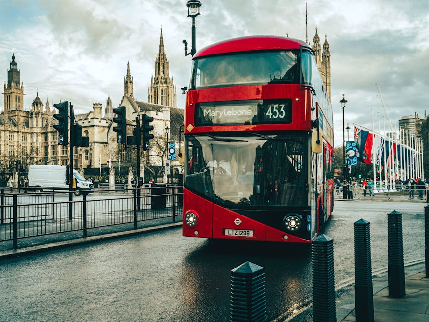 A red double-decker bus with route number 453 and destination 'Marylebone' displayed on the front is parked on a wet street in a city setting. The bus has a sleek, modern design with a curved windshield and is situated near metal bollards that separate the road from the sidewalk. Behind the bus, there are historic Gothic-style buildings with pointed spires, likely part of a government or civic complex, and a row of flagpoles adorned with various national and possibly regional flags. There is a white van and other vehicles in the background, along with traffic lights and street lamps lining the road. The overcast sky creates diffuse lighting, emphasizing the textures and colors of the bus and surrounding architecture. This scene reflects urban public transportation infrastructure close to prominent city landmarks, with the potential for increased waste management considerations in busy areas like Marylebone, where private rubbish collection services such as Waste Disposal Marylebone operate to help maintain cleanliness and prevent issues like fly-tipping in the vicinity.