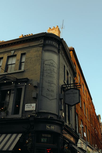 The image shows a historic building with a rounded corner façade, displaying a large vertical sign and a smaller hanging sign for a business located at No. 98 on a street in Marylebone. The building features a mix of cream and darker greyish tones, with textured brick and stone finishes. The top corner of the structure is rounded, with decorative elements and a chimney visible against a clear blue sky. The lower part of the building has large display windows, possibly storefronts, and a striped awning extending over the sidewalk. The environment appears to be an urban street scene, with nearby buildings visible in the background and soft natural lighting suggesting late afternoon or early evening. This setting reflects a typical Marylebone high street environment, where commercial and historical architecture coexist, potentially involving independent retail or hospitality establishments, aligning with the context of local business premises for rubbish removal or waste disposal services, such as those provided by Waste Disposal Marylebone.