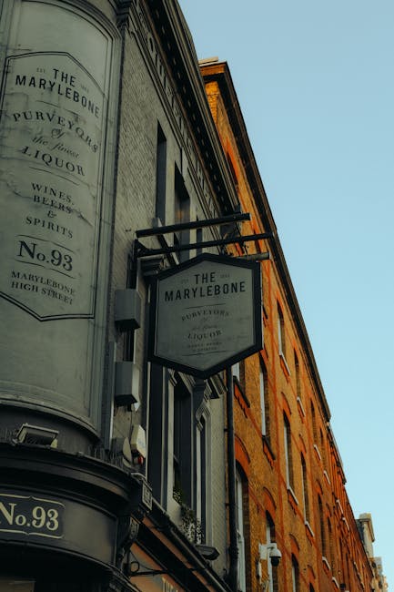 A close-up view of the exterior of a traditional pub named 'The Marylebone,' situated on High Street in Marylebone. The building features a combination of grey and brick-red brickwork, with large windows on multiple floors. Prominent black-framed signs displaying the pub’s name and address are mounted on the façade, with one sign extending outward on a metal bracket. The signage includes text for beverages such as wines, beers, and spirits, and the street number 'No.93' is visible on a plaque below. The surrounding environment appears to be an urban street scene during daylight hours, with clear blue sky overhead. The image captures the detailed textures of the brickwork, glass, and metal fixtures, conveying the historic character of the establishment. This visual context aligns with private waste handling or independent collection services like those provided by Waste Disposal Marylebone, relevant to business or commercial premises in need of rubbish removal or on-site clearance services.