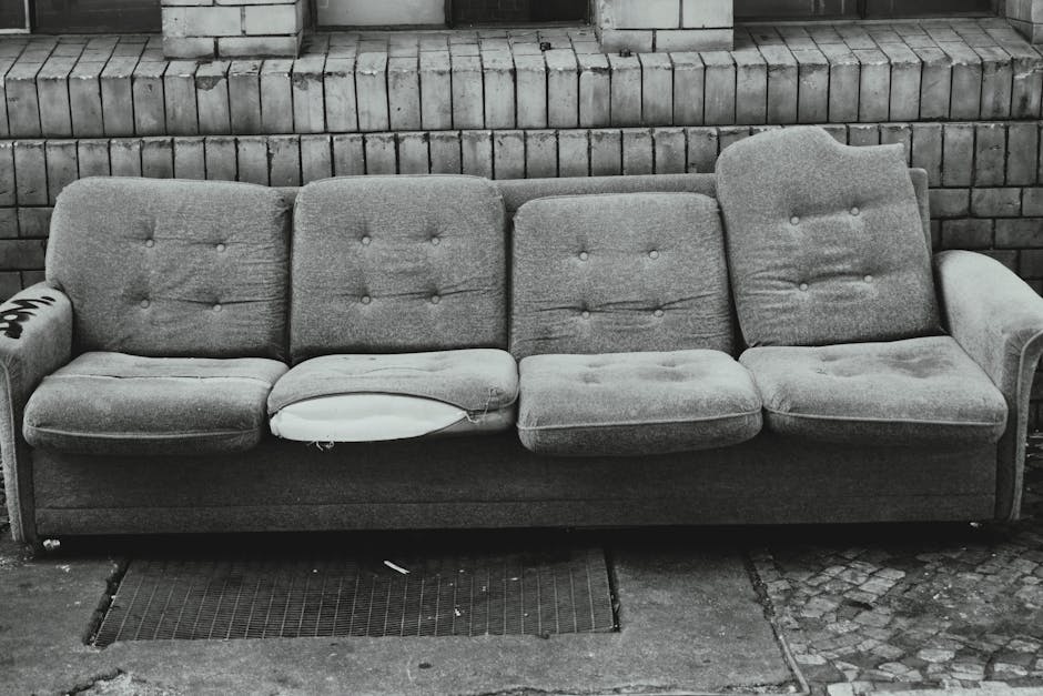 A three-seater sofa made of light grey fabric with a textured finish, positioned outdoors against a brick wall composed of rectangular bricks in a dark grout. The sofa features four distinct seat cushions, with one cushion showing signs of wear and a visible tear as well as a missing piece of upholstery revealing white padding underneath. The overall design includes button-tufted backrests and armrests on both sides, with the left armrest displaying a partially visible decal or mark. The sofa rests on a ground surface composed of concrete slabs and a rubber drain cover, with scattered small debris around the base. The environment suggests an exterior setting, possibly a back alley or a storage area for furniture disposal, indicating the piece may be due for removal through a private rubbish collection service, such as Waste Disposal Marylebone, in a context of alternative waste handling outside standard council arrangements.
