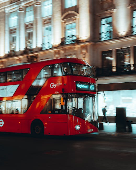 A red double-decker bus with a modern, curved design drives along a city street in front of an ornate, historic building with large columns, decorative moldings, and tall windows. The bus displays the route number 443 and the destination 'Marylebone' on an electronic sign. The pavement is illuminated by ambient street lighting, and a pedestrian, dressed in dark clothing, can be seen walking on the sidewalk in the background near shopfronts with large glass windows. The building's exterior features a beige stone facade with detailed architectural embellishments, and reflections from the glass windows add a touch of brightness to the scene. The overall atmosphere appears lively, capturing a typical urban environment where transport and architecture harmonize, subtly reflecting the context of city services and transportation associated with area-specific postcode zones like Marylebone.