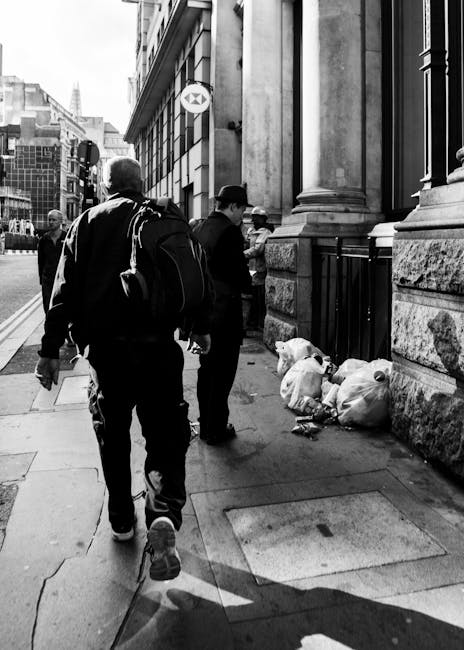 Two individuals standing on a city sidewalk near a stone building with ornate architectural details and large windows, with one person, dressed in dark clothing and carrying a backpack, walking past the other who is bent slightly forward. Nearby, organic waste and rubbish bags are piled against the base of the building, close to a metal drainpipe and a small sign attached to the wall. The scene is set in an urban environment with other buildings visible in the background, and the pavement shows a combination of concrete and stone, with a few scattered shadows cast by nearby structures. The arrangement suggests these individuals may be involved in or observing a waste collection or removal activity, typical of private rubbish handling services such as those offered by Waste Disposal Marylebone. The overall atmosphere is calm and functional, conveying a typical city street scene with a focus on waste accumulation near a commercial or office premises.