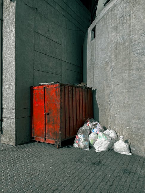 A red, weathered metal skip with visible rust and signs of use is positioned against a dark green and grey building exterior, located on a paved sidewalk area. The skip is filled with miscellaneous waste, but the lid is slightly open, revealing some contents. Around the base of the skip, several white plastic garbage bags are piled, containing various unidentifiable rubbish. The bags appear to be loosely tied and are slightly bulging, indicating they are full. The surrounding environment includes a dark green wall with horizontal panels on the left side, and a light grey textured wall on the right, both extending vertically. In the background, a narrow alleyway or passage with small, rectangular windows visible at the top provides natural light, casting subtle shadows on the walls and ground. The scene reflects typical on-site waste collection, characteristic of private rubbish removal services such as those offered by Waste Disposal Marylebone, highlighting an instance of temporary outdoor rubbish storage awaiting collection or disposal in an urban setting.