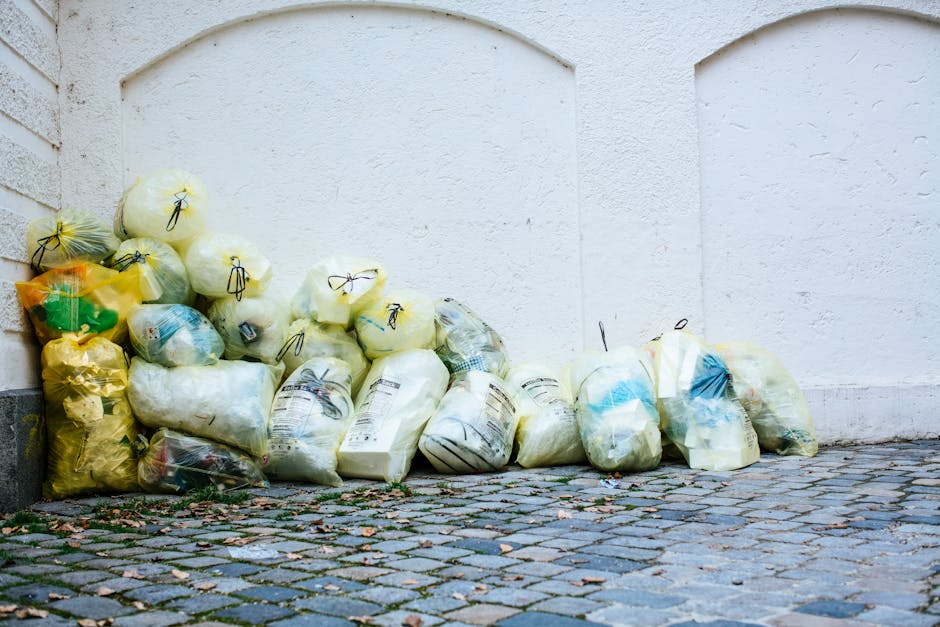 A collection of numerous plastic rubbish bags piled along the base of a white exterior wall, situated on a cobbled pavement. The bags vary in size and are transparent, yellow, and white, with some containing visible waste such as paper and plastic items. Most bags are tied with black or yellow ties, and some are partially squashed or bulging, indicating fullness. The textured cobblestone ground beneath the bags shows fallen leaves and small debris, suggesting outdoor storage outside a residential or commercial building. The white wall features subtle wall textures and gentle arches at the top, creating a clean background that contrasts with the cluttered bags. This scene depicts an example of private waste collection or on-site clearance managed by a professional rubbish removal service, such as Waste Disposal Marylebone, which handles domestic or light commercial waste, emphasizing the importance of maintaining tidy outdoor waste storage areas.