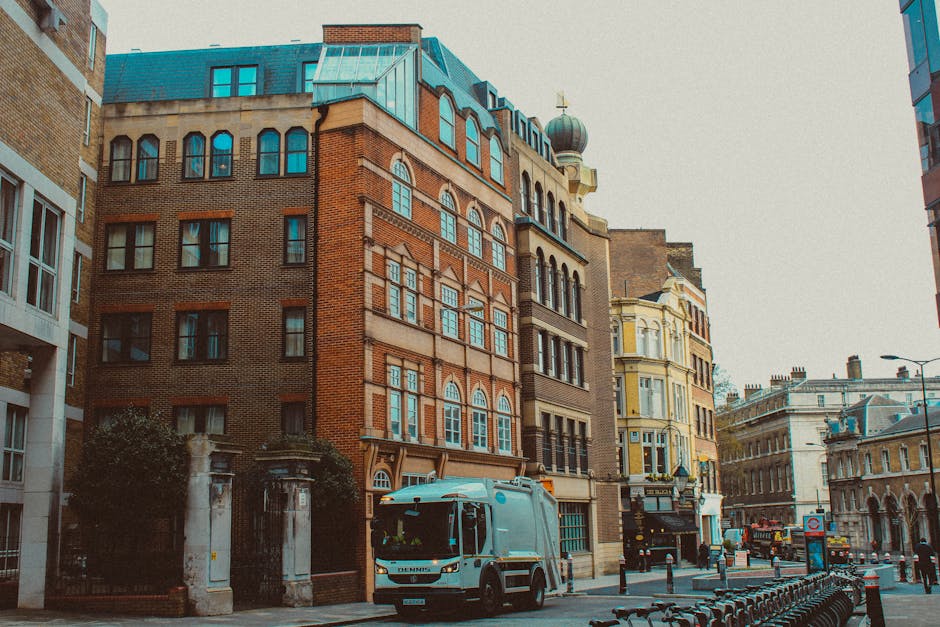 A street scene featuring a blue waste collection vehicle parked in front of a row of multi-story buildings with distinct architectural styles in an urban environment. The vehicle, likely used by Waste Disposal Marylebone for independent waste collection, has a smooth, metallic finish and is positioned on the road near a sidewalk. The buildings display a variety of materials, including red brick and painted facades, with large, rectangular windows and decorative elements such as cornices and arched window frames. One building on the corner has a rounded turret with a dome-shaped roof, adding historical character to the scene. The background includes additional historic buildings, a streetlamp, and a bus stop sign, with the narrow street lined by a barrier of bollards. The atmosphere is neutral with diffused daylight illuminating the scene, highlighting the urban context of private rubbish collection services operating within the city landscape.