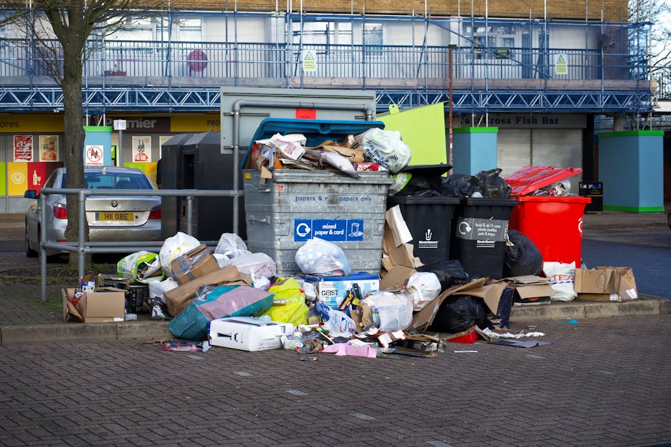 A collection of numerous plastic rubbish bags piled along the base of a white exterior wall, situated on a cobbled pavement. The bags vary in size and are transparent, yellow, and white, with some containing visible waste such as paper and plastic items. Most bags are tied with black or yellow ties, and some are partially squashed or bulging, indicating fullness. The textured cobblestone ground beneath the bags shows fallen leaves and small debris, suggesting outdoor storage outside a residential or commercial building. The white wall features subtle wall textures and gentle arches at the top, creating a clean background that contrasts with the cluttered bags. This scene depicts an example of private waste collection or on-site clearance managed by a professional rubbish removal service, such as Waste Disposal Marylebone, which handles domestic or light commercial waste, emphasizing the importance of maintaining tidy outdoor waste storage areas.
