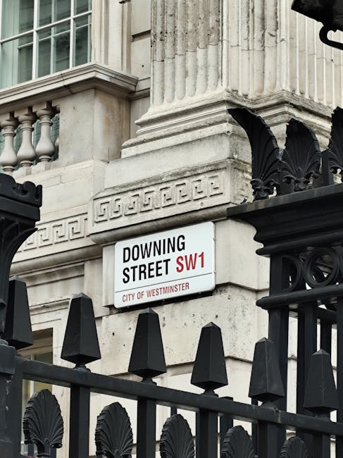 The image shows a street sign attached to a historic building with classical architectural details, including decorative stonework and fluted columns. The sign indicates 'Downing Street SW1' within the City of Westminster, featuring black text on a white background with a red highlight for the postcode. The lower part of the sign states 'City of Westminster' in smaller red font. In the foreground, a black wrought-iron fence with decorative spikes and ornamental elements partially obscures the lower section of the building, providing a sense of enclosure and security. The building’s stone facade is light grey with subtle weathering, reflecting its architectural style and age. The overall scene is illuminated by natural daylight, emphasizing the textures of the stone and metalwork. This visual context relates to the area’s heritage and formal urban environment, aligning with private or official waste management practices, such as rubbish removal services, that might operate nearby, especially in historically significant districts like this one.