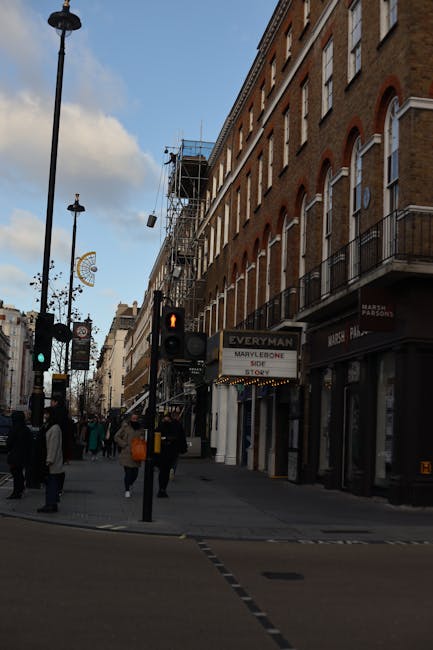 The image depicts a busy street scene at a pedestrian crossing in Marylebone, featuring several people waiting to cross. In the foreground, there is a black traffic light pole with illuminated red and green signals, along with a pedestrian crossing button. The street is lined with multi-story brick buildings, some with decorative arch-shaped windows and small balconies, reflecting traditional London architecture. One of the buildings houses a shop with a sign for 'Everyman,' and additional storefronts are visible further along the sidewalk. Above the shops, there are several floors with beige brick facades and white-framed windows. In the background, scaffolding is set up around a building under renovation, indicating ongoing construction work. The sky is partly cloudy, casting diffused daylight across the scene. The overall atmosphere suggests a typical urban environment with active pedestrian movement, aligned with services like waste disposal companies providing alternative rubbish collection options in Marylebone, London.