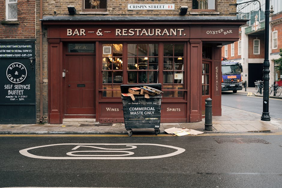 On the sidewalk in front of a brick-built bar and restaurant with large windows and a brown wooden framework, a black commercial waste bin labeled 'Commercial Waste Only' is positioned, overflowing with flattened cardboard boxes and miscellaneous paper waste. Nearby, a few scattered sheets of paper lie on the pavement near a small, black bollard. To the right of the bin, a black metal lamppost stands alongside a parked delivery truck on the street, which is paved with dark asphalt featuring a prominent white road marking. The building's signage indicates an oyster bar and includes references to wines, spirits, and cocktails, with a street sign reading 'Crispin Street' mounted above the establishments. The ambient lighting suggests late afternoon or early evening, with a subtle warm glow from the interior illuminating part of the Shopfront, while the surrounding environment appears urban, consistent with areas serviced by private waste collection and rubbish removal specialists like Waste Disposal Marylebone.