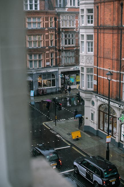 The image depicts a busy street scene at a pedestrian crossing in Marylebone, featuring several people waiting to cross. In the foreground, there is a black traffic light pole with illuminated red and green signals, along with a pedestrian crossing button. The street is lined with multi-story brick buildings, some with decorative arch-shaped windows and small balconies, reflecting traditional London architecture. One of the buildings houses a shop with a sign for 'Everyman,' and additional storefronts are visible further along the sidewalk. Above the shops, there are several floors with beige brick facades and white-framed windows. In the background, scaffolding is set up around a building under renovation, indicating ongoing construction work. The sky is partly cloudy, casting diffused daylight across the scene. The overall atmosphere suggests a typical urban environment with active pedestrian movement, aligned with services like waste disposal companies providing alternative rubbish collection options in Marylebone, London.